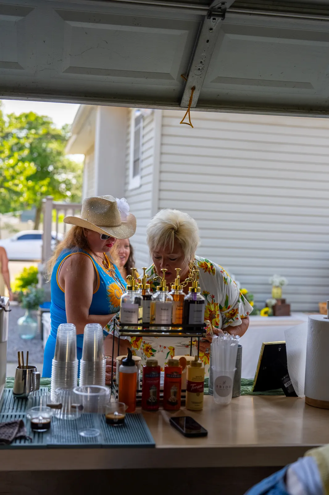 Two guests in summer outfits read the syrup labels at the Abide Coffee Co. cart beneath an open garage door.