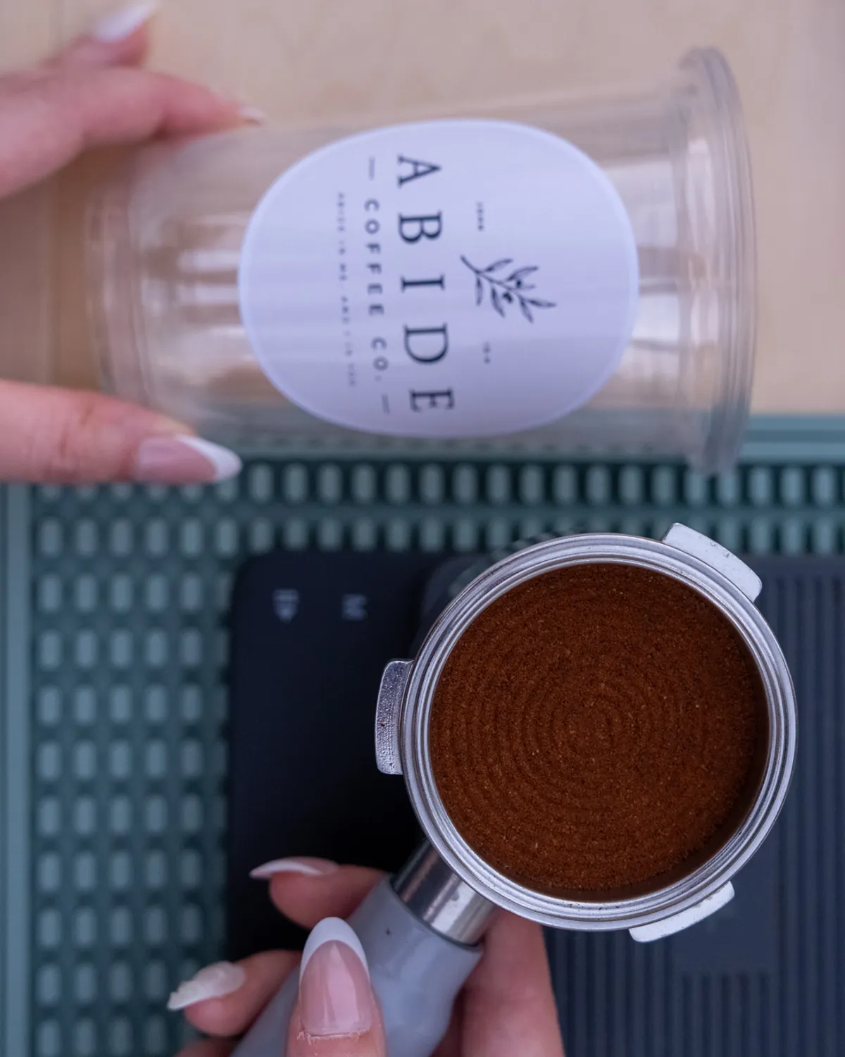 Close overhead view of a hand holding a clear Abide Coffee Co. cup above a portafilter of freshly tamped espresso.