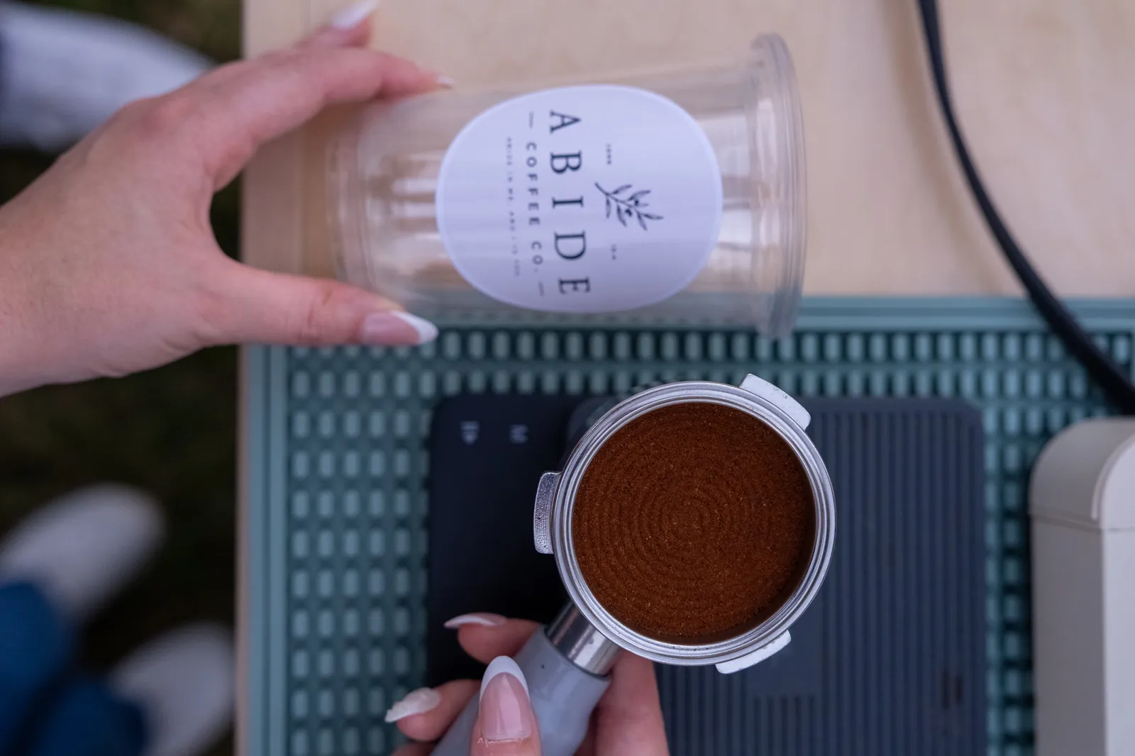 Overhead view of a hand setting a clear Abide Coffee Co. cup beside a tamped portafilter, a guest's shoes on the grass below.